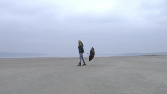 Pretty young girl pulling umbrella against wind walking from right to left on sandy beach. Quiet weather, calm water, fog, mist and haze over water. Fantastic milky landscape movie film.