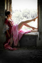 Beautiful Young Woman in Victorian Dress sitting on the Window in a Dark Room. Fairy Tale. Sun Light Effect