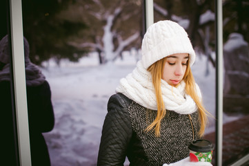 woman in white scarf and hat drinks coffee. cute young blond woman standing near mirror reflection showcases cafe, drinking coffee to go, holding Christmas paper cup in his hands