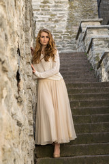 Fototapeta na wymiar Caucasian white female model and brick stone. Beautiful girl, long red hair, beige skirt and cardigan. Woman standing on the stairs in old town, limestone walls background