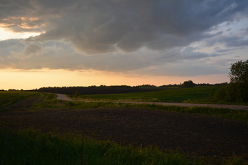 Obraz premium piękny wiosenny wiejski zachód słońca nad polem, oddzielony piaszczystą drogą, przyrodą, krajobrazem, jasnym niebem i ciemnymi chmurami