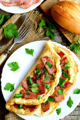 Delicious omelette on a plate. Omelette stuffed with fried bacon and decorated with chopped fresh parsley. Sliced bacon, bread, cutlery, fresh parsley leaves on wooden background. Top view. Closeup © onlynuta