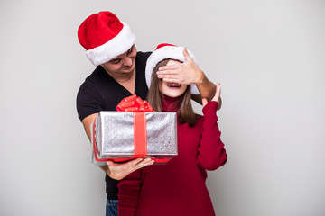 Handsome young man holding a gift box while her girlfriend standing behind him and covering his eyes with hands and wearing santa hat christmas