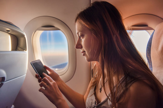 Young woman using mobile phone in airplane at sunset