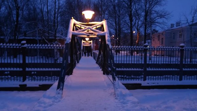 Winter night over the bridge into the distance are people, the bridge lights a lantern in the background trees and the right of the building, from the bridge to the right and left cast-iron fence.