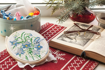 Winter still life : hoop with embroidered flowers pattern, open book, glasses, teapot and cup, colorful yarn in box and christmas tree in vase. Selective focus. Home decor, relax concept