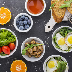 Breakfast on a dark background: fried quail eggs, arugula, fresh bread, lettuce, tomatoes, oranges, nuts, tea, coffee,  mushrooms,  blueberries. Top view