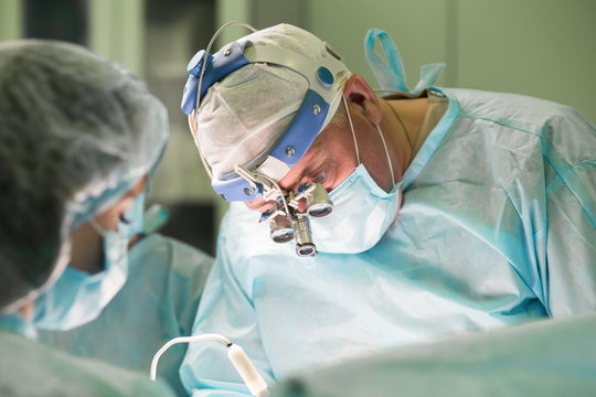 Surgeon and his assistant performing cosmetic surgery in hospital operating room. Surgeon in mask wearing loupes during medical procedure. Breast augmentation, enlargement, enhancement