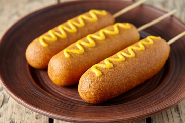 American corn dog street junk food deep fried hotdog meat sausage with mustard snack treat coated in a thick layer of cornmeal batter on stick unhealthy eating on rustic wooden table.