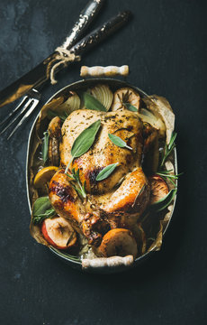 Oven roasted whole chicken with onion, apples and sage in serving tray with cutlery over dark stone background, top view, selective focus. Celebration food concept