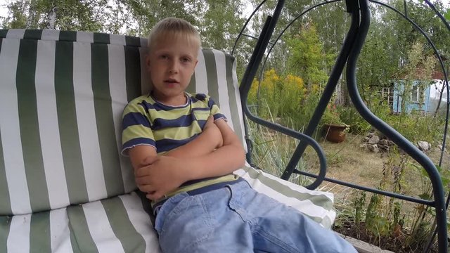 Little blond boy (7 years old) says, swinging on the swing in the summer nature 