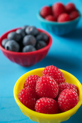 Berries in colorful bowls lined up on blue background, viewed from an angle