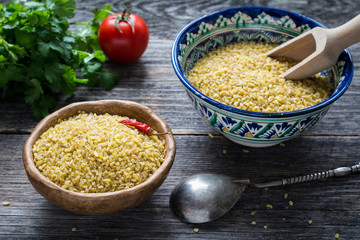 Raw bulgur wheat grains in colorful arabic bowl, fresh parsley, tomato and peppers for cooking. Wooden table background, horizontal composition