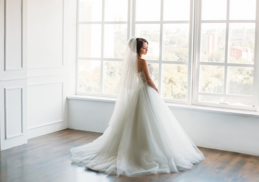 Beautiful brunette girl standing in a bright room.She is dressed in luxurious, fluffy, white dress.Gentle puppet's face.European image of bride.Photo high key style.Fashionable toning.Creative color.