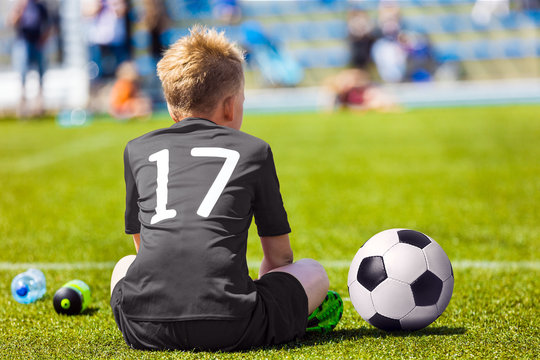 Child in black sportswear with soccer ball. Boy sitting on football pitch. School football match for children
