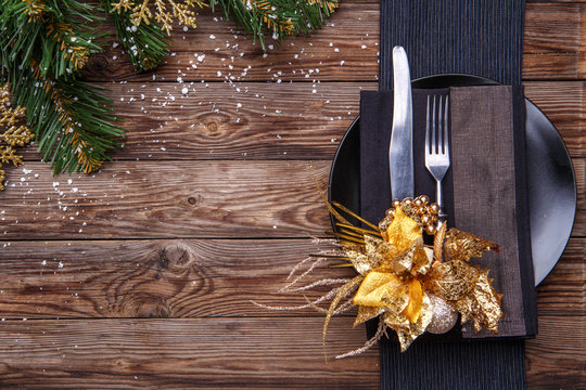 Christmas table place setting with black napkin, plate, fork and knife, decorated gold flower and christmas pine branches.