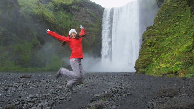 Happy woman jumping excited by waterfall on Iceland waving hand saying hello by Skogafoss. Girl woman looking at camera cheerful and joyful visiting tourist attraction landmarks in Icelandic nature.