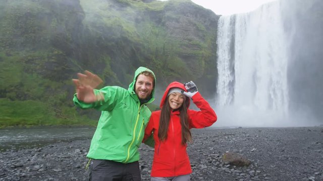 People by Skogafoss waterfall on Iceland waving hands saying hello having fun on golden circle. Couple visiting famous tourist attractions and landmarks in Icelandic nature landscape.