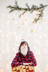 A brunette girl in a Christmas Santa Claus costume, with a beard and hat, in a red checkered shirt, crawled out of a gift box, laughing.In the background a blurred branch of a tree with lights and toy