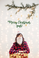 A brunette girl in a Christmas Santa Claus costume, with a beard and hat, in a red checkered shirt, crawled out of a gift box, laughing.In the background a blurred branch of a tree with lights and toy