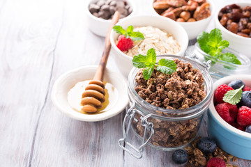 Homemade chocolate granola ingredients, nuts, oats, honey and berries on white wooden background. Healthy breakfast concept with copy space.