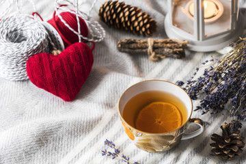 cup of hot tea with lemon on wool shawl with  burning candle, cones, knitted red heart and lavender