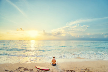 Fototapeta na wymiar Surfing and meditation. Enjoying sunset.. Relaxed young man sitting on lotus position with surf board on the beach.