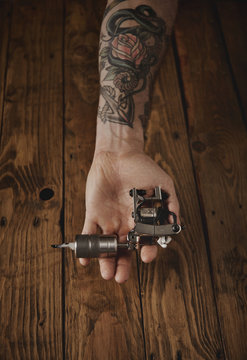 Close up of a man's hand offers new custom made shiny metal tattoo gun on camera, above rustic wooden table
