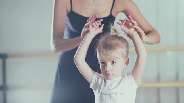 Cute little girl in the classroom with the teacher of ballet. Girl in a white dance form and dance teacher in a black suit. Little girl raises her arms in the third position. Children's ballet school.