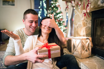 Couple in front of fireplace. Man givin present to woman.