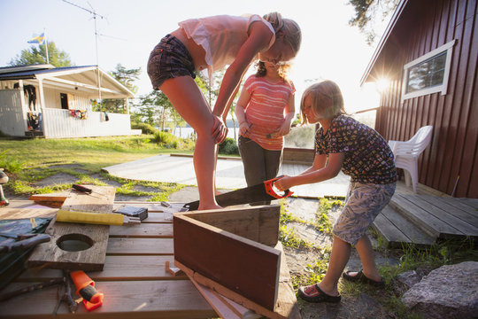Girls looking at brother sawing wooden plank in yard