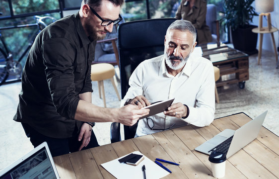 Concept of coworking people.Young bearded man showing startup idea on mobile tablet.Adult businessman working together at the wooden table with partner.Horizontal,blurred.