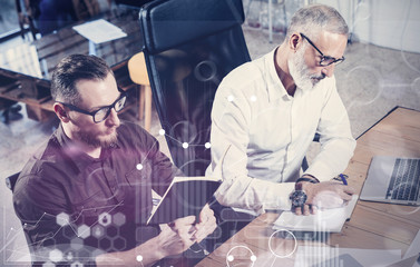Concept of digital screen,virtual connection icon,diagram, graph interfaces.Bearded young man working together with adult business partner at the wooden table.Coworkers teamwork in modern office.Flare