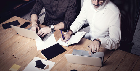 Concept of business teamwork.Two bearded businessmans discussing ideas together in modern office.Adult man making notes in paper notebook,using gadgets at wooden table.Horizontal,film effect.