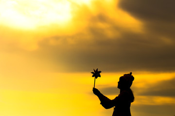 Fototapeta na wymiar Silhouette of Beautiful girl holding wind toy or wind turbine or pinwheel and wool hat with golden sky on winter season in morning.