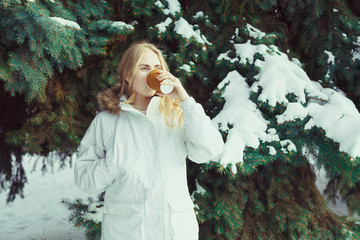 young blond woman in a white jacket drinking coffee on a background of snow-covered trees