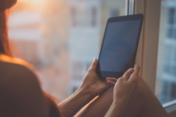 Woman using digital tablet at home sitting near window.