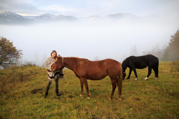Beautiful girl with horses at mountain peak above clouds and fog. Girl with an horse playing together at the farm. Young woman take care of her horse. Early morning.Misty mountain.