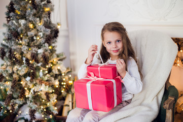 Obraz na płótnie Canvas Beautiful girl near Christmas tree unpacking presents sitting on a chair