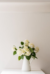 White hydrangeas in jug on table against white wall with vintage picture rail (selective focus)