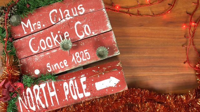 Endless looping video of overhead Christmas table top with a vintage sign arrow pointing to the North Pole and Mrs. Claus cookies company. Room for text on wooden surface on the right side.