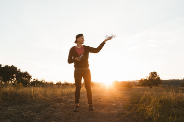 Young man standing with drone outdoor, sunset flare. Presentation of unmanned aerial vehicle at nature, successful landing to hand by remote controller