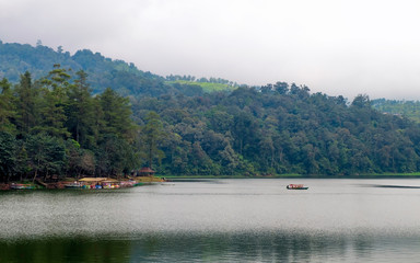 Beautiful scenery of huge lake, with trees, and mist create calming atmosphere. In the middle of the lake, there also a little boat departing from ports, going into the middle of lake