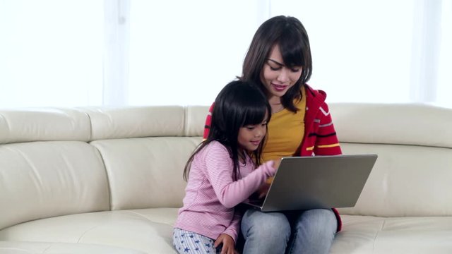 Young Asian mother and her daughter sitting on the sofa while using a laptop computer together