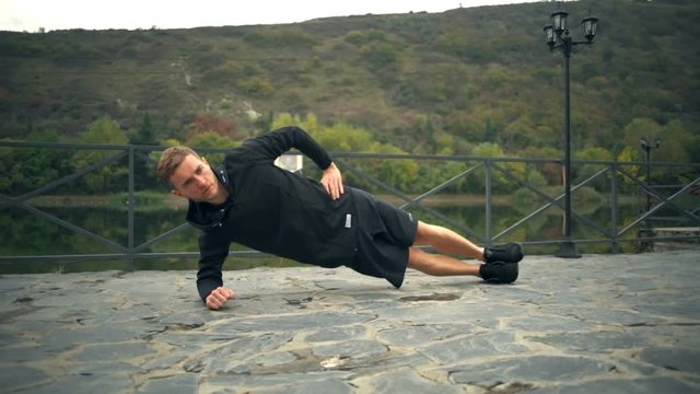 A young handsome man doing side plank touching ground on a terrace near the pond