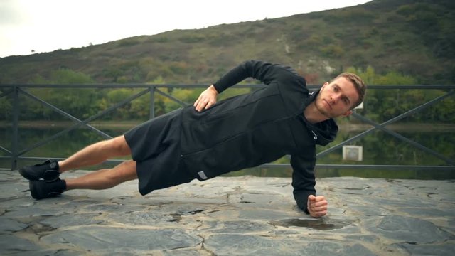 A young handsome man doing side plank touching ground on a terrace near the pond