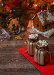Two glasses of cocoa with marshmallows, whipped cream and chocolate syrup on wooden table and Gingerbread house, cookie jar and christmas tree with toys and garland on background.