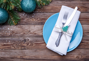 Christmas table place setting with blue plate, fork and knife, decorated ribbon and bow with fir-tree toy, white napkin and christmas pine branches with christmas balls.