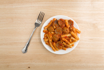 Rigatoni pasta with meatballs and sausage on plate with fork atop a wood table.