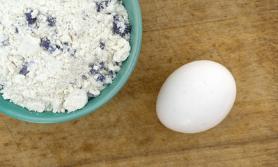 Bowl of blueberry muffin mix with an egg atop an antique wood cutting board.